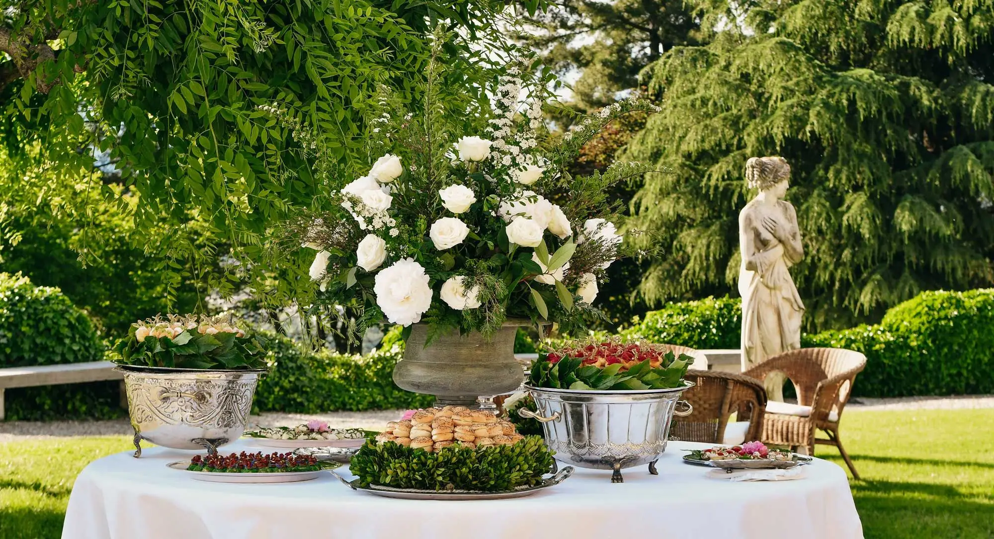 Un ricevimento in giardino: tavoli bianchi, fiori e alzate d'argento fra gli alberi